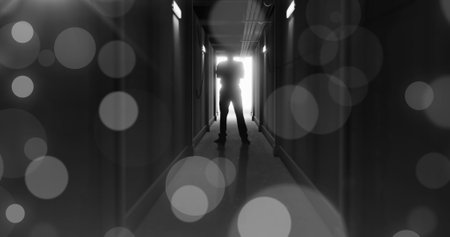 Image of grey bokeh light spots over scary figure backlit in doorway at end of dark corridor. Halloween, fear, horror and suspense concept digitally generated image.の写真素材