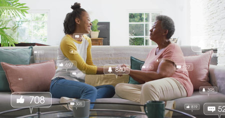 Holding hands mother and daughter sitting on gray sofa in living room, with social media icons....