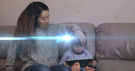 Image of light moving over happy asian mother and son with tablet sitting on sofa. Family, spending quality time together concept digitally generated image.の写真素材