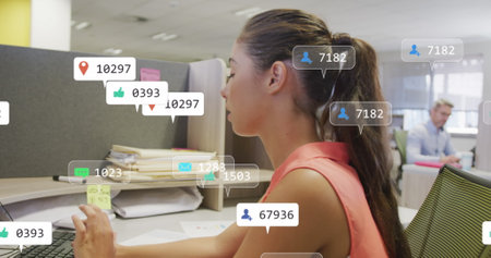 Image of social media notifications processing data over caucasian woman working at office desk. Global communication network, digital interface and social media concept digitally generated image.の写真素材