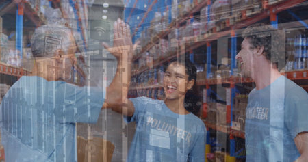 Hispanic woman wearing volunteer shirt high-fiving colleague in warehouse with metal shelves, boxes. Collaboration, teamwork, community, industrial, candid, team spirit, productivityの写真素材