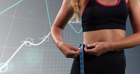 Wrapping woman in sportswear measuring waist with blue measuring tape in gym, with data graphs. Athletic, fitness, discipline, determination, health, technology, modernの写真素材