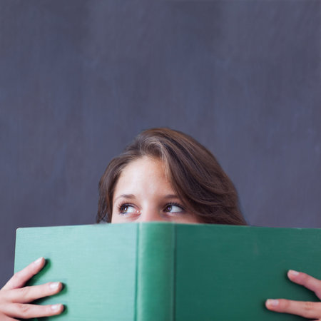 Woman is peeking over green hardcover book in flat design with smooth grey backdrop, copy space. Portrait, curiosity, literary, minimalism, contemplative, interior, expressionの写真素材