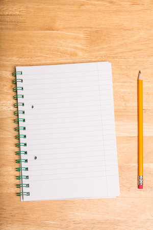 Spiral-bound lined notebook is lying open on wooden tabletop, with yellow pencil resting parallel....