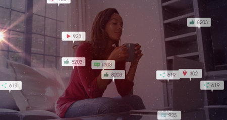 Woman holding gray mug and checking silver laptop in living room with social media icons. Cozy, modern, technological, casual, relaxing, communication, workの写真素材