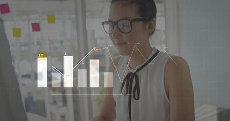 Reviewing charts businesswoman wearing glasses at office computer with glass partition notes....