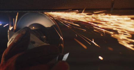 Cutting steel pipe with gloved hand holding angle grinder in dim workshop, with orange sparks. Industrial, craftsmanship, precision, safety, metallic, rugged, workshopの写真素材