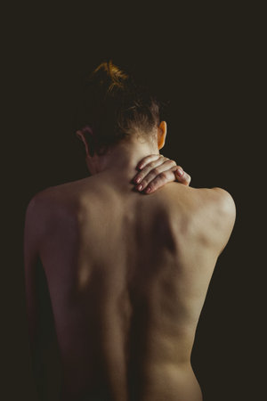 Woman is showing bare back in dark studio portrait, highlighting spine ridges and bun. Minimalist, elegance, contemplative, moody, silhouette, artistic, wellnessの写真素材