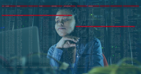 Mid adult blue-shirted woman studying code on glass panel at office, with monitors and keyboard....