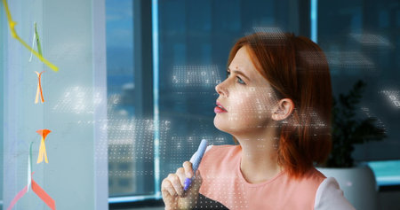 Woman analyzing diagrams, data on glass board at office window, with blue marker and potted plant....