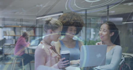 Collaborating three women standing around tall table in workspace, with laptop cup bookshelves bike....