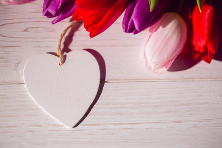 Flat design white wooden heart ornament is resting on tabletop with red, pink, purple tulip bouquet. Romantic, rustic, minimalist, wedding, decor, spring, naturalの写真素材