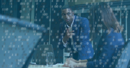 Gesturing businessman in dark blue suit leaning on conference table at office, with laptop and code....