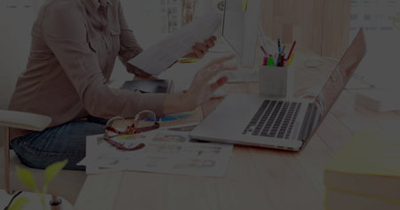 Woman wearing button-down shirt holding documents while using laptop at office desk, with pencils....