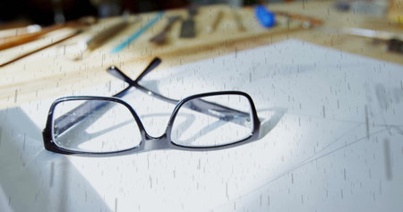 Resting black rectangular eyeglasses open on white paper at workspace, with blurred drawing tools. Minimalist, creative, workspace, design, intellectual, artistic, precisionの写真素材