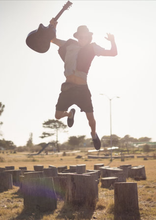 Man holding guitar jumping on tree stumps during sunny day