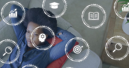 Reclining woman in denim shirt on gray sofa at home, with red pillow and digital icons. Relaxation,...