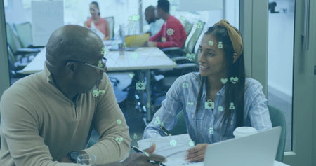 Collaborating man wearing glasses and woman writing on laptop at table, with digital health icons....
