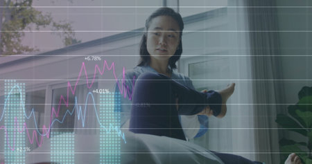 Stretching Korean woman in navy leggings, grey top on mat by window, floating financial graphs. Wellness, mindfulness, fitness, home office, modern, productivity, relaxationの写真素材