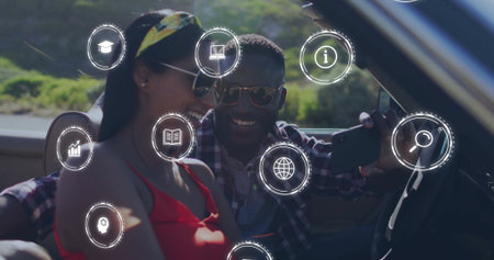 Smiling couple wearing sunglasses using phone in convertible on hillside road, with digital icons....