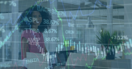 Smiling businesswoman holding laptop in modern open-plan office, with stock market chart overlays....