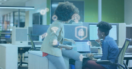 Examining security icons on laptops, two professionals leaning over desk in modern open-plan office....