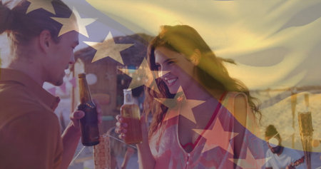 Toasting couple holding beer bottles clinking near beach cabana at sunset, with tiki torch....