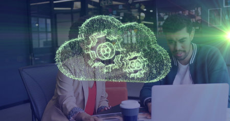 Tech colleagues collaborating at office with laptop cup, docs and holographic cloud icon with gears....