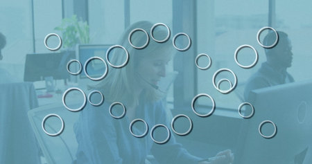 Typing on tablet, woman in sweater assisting customer in open-plan office, with headset microphones. Collaboration, communication, teamwork, modern, professional, technology, productivityの写真素材