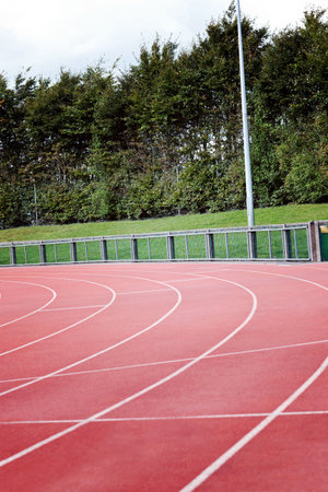 Curved red track is winding in flat design with white lanes, railing, hedge and light pole....