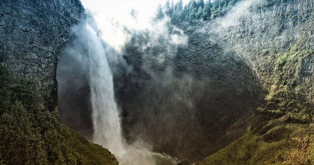 Narrow waterfall is plunging down rugged cliff in flat design, with mist, moss, evergreen trees. Nature, adventure, landscape, tranquil, wilderness, scenery, remotenessの写真素材