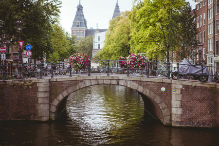 Stone arch canal bridge is spanning canal in flat style, with bicycles and hanging flower baskets. Architectural, urban, picturesque, floral, vintage, scenic, tranquilの写真素材