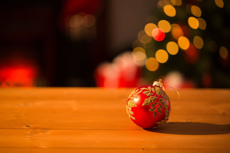 Red bauble with gold leaf is resting on wooden tabletop in 3D render, glowing under bokeh. Festive, luxury, shimmering, cozy, decorative, celebration, warmの写真素材