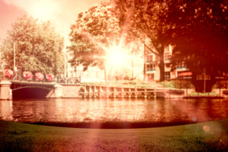 Flat design sunlit arched stone bridge is spanning canal by grassy bank, flower baskets, sun flare. Urban, landscape, nature, tranquility, architecture, reflection, foliageの写真素材