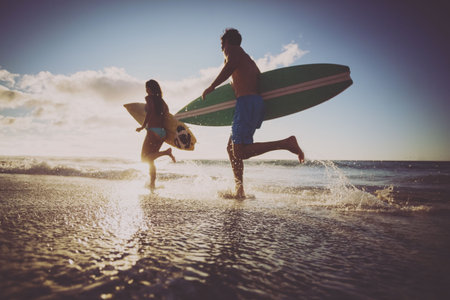 Low angle view of cheerful caucasian couple with surfboards running in sea against sky at sunset. Unaltered, love, together, vacation, enjoyment and nature conceptの写真素材