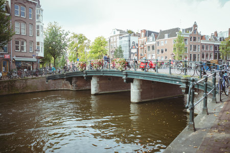Flat design is depicting green metal pedestrian bridge spanning brown canal with flower baskets. Urban, scenic, vibrant, historic, cycling, leisure, natureの写真素材