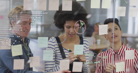 Collaborating team placing pastel sticky notes on glass wall in office with pen, marker diagrams....