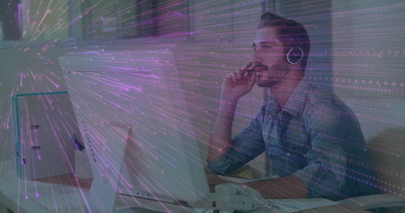 IT pro wearing blue shirt thinking in office with two monitors, keyboard, handset, mug, headphones....