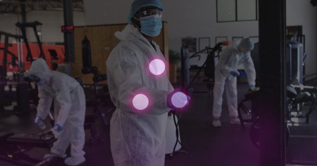 Sanitizing male worker wearing protective coverall disinfecting machines at gym, with spray wand....