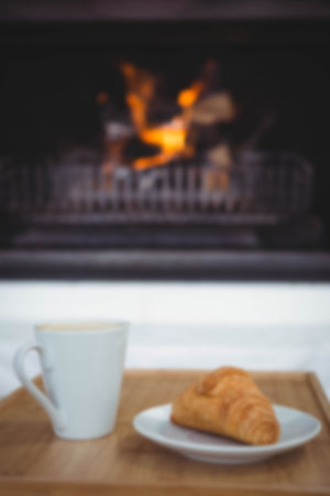 Coffee cup by croissant in plate on table against fireplace at home