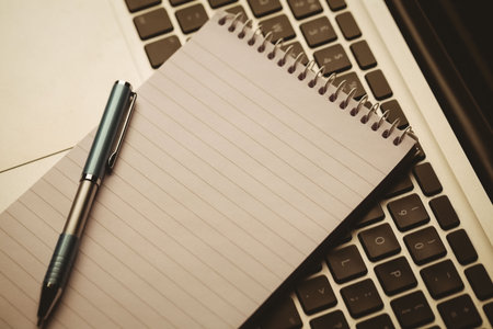 Spiral-bound lined notebook is resting on laptop keyboard in flat design, showing blue-grip pen....