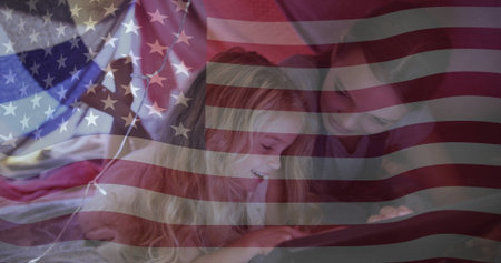 Lying sisters wearing pajamas focusing on tablet on bed, with blankets, pillows and flag overlay. Family, children, bonding, leisure, relaxation, cozy, patrioticの写真素材