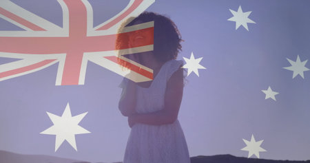Standing woman wearing white lace dress on dawn hills, with translucent Australian flag overlay. Bohemian, patriotic, vibrant, ethereal, contemplative, scenic, spiritualの写真素材