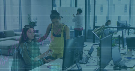 Pointing woman in yellow overalls guiding seated coworker using dual monitors at open-plan office....