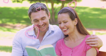 Reading pair wearing pink top, blue shirt sharing light green book on wooden park bench. Romance, intimacy, leisure, outdoor, tranquility, connection, natureの写真素材