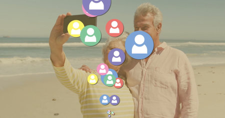 Taking selfie older couple standing on sandy beach, with smartphone and floating social media icons. Senior, companionship, outdoor, leisure, digital, connectivity, lifestyleの写真素材