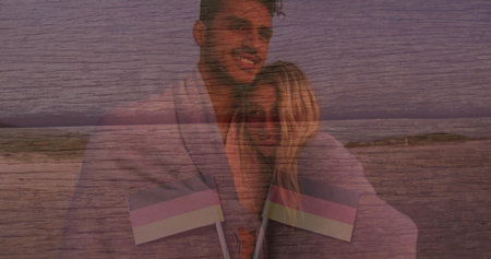 Smiling couple wearing light-colored blanket on sandy beach at dusk, holding small handheld flags....