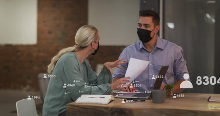 Masked colleagues reviewing papers at office desk, with laptop, plant and digital overlay. Collaboration, teamwork, modern, professional, productivity, data, communicationの写真素材