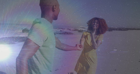 Holding hands couple wearing green T-shirt and yellow sundress smiling on beach, with ocean waves....