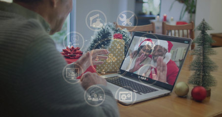 Senior man in sweater waving at laptop during call at dining table, with interface icons. Festive,...